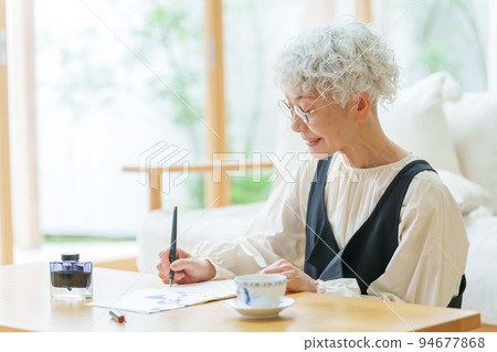Gray-haired woman writing a letter in the living room 94677868