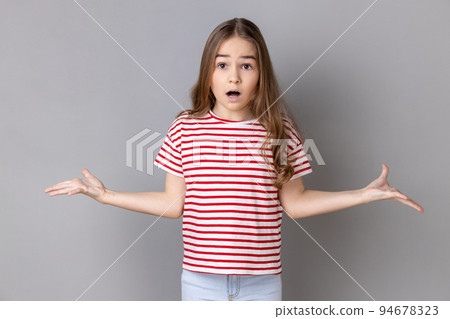What do you want. Portrait of little girl wearing striped T-shirt standing with raised hands and surprised indignant expression, asking what reason. Indoor studio shot isolated on gray background. 94678323