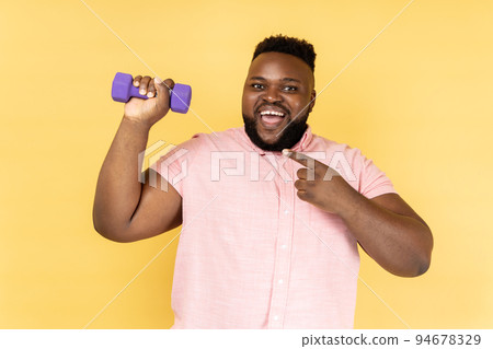 Portrait of excited positive bearded man wearing pink shirt standing with dumbbell and pointing at sport equipment, looking at camera. Indoor studio shot isolated on yellow background. Portrait of excited positive bearded man wearing pink shirt standing with dumbbell and pointing at sport equipment, looking at camera. Indoor studio shot isolated on yellow background. 94678329