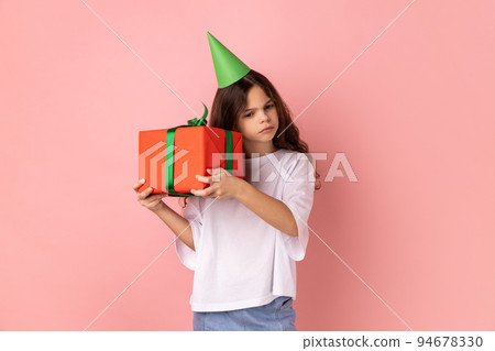 Portrait of curious little girl wearing white T-shirt and party cone holding red wrapped present box, being interested white inside of gift. Indoor studio shot isolated on pink background. 94678330