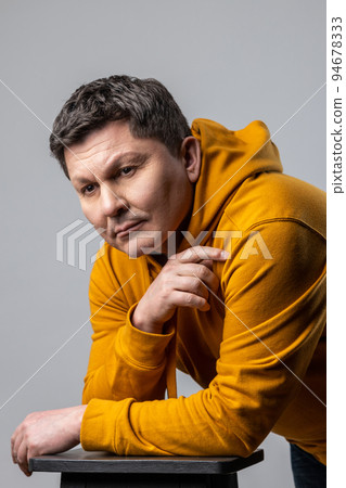Portrait of handsome middle aged man looking pensive thoughtful, leaning on chair, being deep in thoughts, wearing urban style hoodie. Indoor studio shot isolated on gray background. Portrait of handsome middle aged man looking pensive thoughtful, leaning on chair, being deep in thoughts, wearing urban style hoodie. Indoor studio shot isolated on gray background. 94678333