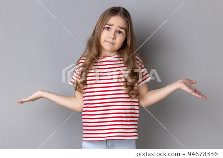 I don't know. Portrait of ambiguous little girl wearing striped T-shirt shrugging shoulders with no idea gesture, clueless embarrassed face. Indoor studio shot isolated on gray background. 94678336