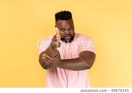I'll kill you. Portrait of man wearing pink shirt pointing finger guns to camera and looking aggressive, threatening to shoot, hands imitating weapon. Indoor studio shot isolated on yellow background. 94678345