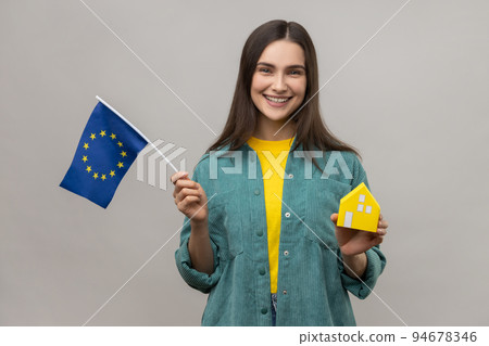 Portrait of positive satisfied holding european flag and paper house, dreaming to buy accommodation in Europe, wearing casual style jacket. Indoor studio shot isolated on gray background. Portrait of positive satisfied holding european flag and paper house, dreaming to buy accommodation in Europe, wearing casual style jacket. Indoor studio shot isolated on gray background. 94678346