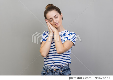 Portrait of sleepy bored woman wearing striped T-shirt leaning head on hands, keeps eyes closed, having nap, feeling tired and exhausted. Indoor studio shot isolated on gray background. Portrait of sleepy bored woman wearing striped T-shirt leaning head on hands, keeps eyes closed, having nap, feeling tired and exhausted. Indoor studio shot isolated on gray background. 94678367