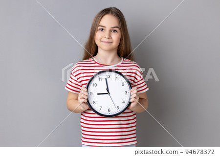 Portrait of smiling little girl wearing striped T-shirt holding big wall clock, looking at camera with pleasant emotions, time to go. Indoor studio shot isolated on gray background. 94678372