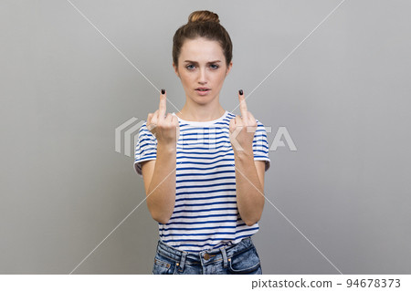 Portrait of crazy angry woman wearing striped T-shirt showing middle finger, rude gesture, face full of hatred and resentment. Indoor studio shot isolated on gray background. 94678373