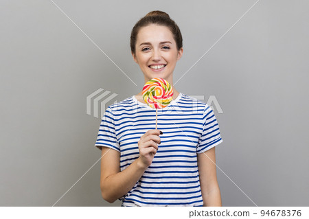 Portrait of smiling woman wearing striped T-shirt holding striped colorful lollipop on stick in her hand, crazy about sweet confectionery. Indoor studio shot isolated on gray background. 94678376