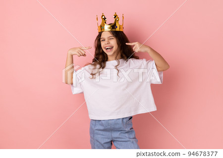 Portrait of happy confident smiling little girl wearing white T-shirt standing in gold crown, pointing at herself with pride, looking at camera. Indoor studio shot isolated on pink background. Portrait of happy confident smiling little girl wearing white T-shirt standing in gold crown, pointing at herself with pride, looking at camera. Indoor studio shot isolated on pink background. 94678377