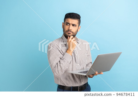 Portrait thoughtful pensive man with beard holding laptop and holding chin looking away, thinking over new business project, wearing striped shirt. Indoor studio shot isolated on blue background. Portrait thoughtful pensive man with beard holding laptop and holding chin looking away, thinking over new business project, wearing striped shirt. Indoor studio shot isolated on blue background. 94678411