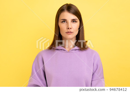 Portrait of young serious unsmiling strict woman looking at camera with calm attentive focused expression, wearing purple hoodie. Indoor studio shot isolated on yellow background. Portrait of young serious unsmiling strict woman looking at camera with calm attentive focused expression, wearing purple hoodie. Indoor studio shot isolated on yellow background. 94678412