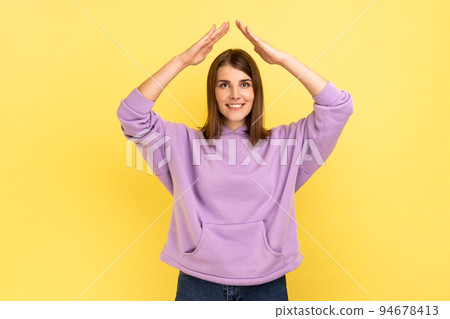 I'm in safety. Portrait of happy protected woman raising hands above head, making roof gesture, insurance, wearing purple hoodie. Indoor studio shot isolated on yellow background. 94678413