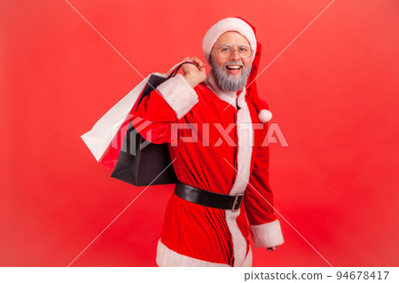 Portrait of smiling elderly man with gray beard wearing santa claus costume standing with shopping bags, buying present for winter holidays. Indoor studio shot isolated on red background. 94678417
