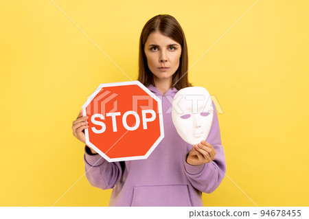 Portrait of serious woman holding red stop sign and white mask, looking at camera with strict bossy expression, wearing purple hoodie. Indoor studio shot isolated on yellow background. 94678455