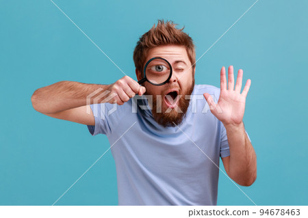 Portrait of funny bearded man holding magnifying glass and looking at camera with big zoom eye, curious face, waving hand, saying hi. Indoor studio shot isolated on blue background. 94678463