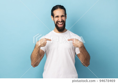 Portrait of delighted man with beard wearing white T-shirt pointing fingers on himself and looking at camera with toothy smile, extremely satisfied. Indoor studio shot isolated on blue background. Portrait of delighted man with beard wearing white T-shirt pointing fingers on himself and looking at camera with toothy smile, extremely satisfied. Indoor studio shot isolated on blue background. 94678469