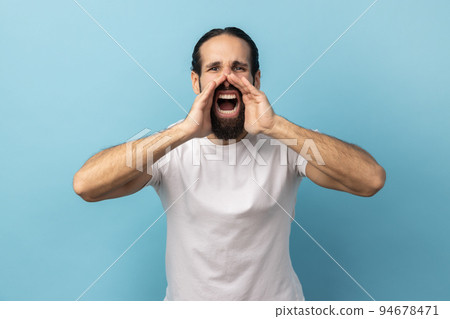 Attention, announcement. Portrait of handsome man with beard wearing white T-shirt screaming message, loud advertising, keeps hands near mouth. Indoor studio shot isolated on blue background. Attention, announcement. Portrait of handsome man with beard wearing white T-shirt screaming message, loud advertising, keeps hands near mouth. Indoor studio shot isolated on blue background. 94678471