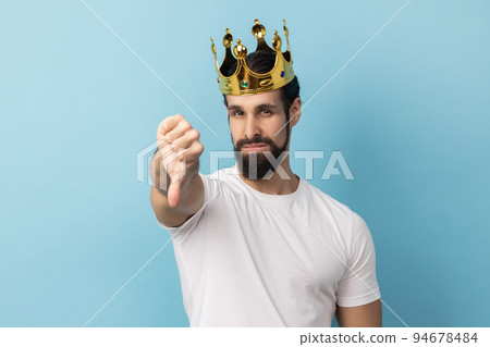 Portrait of sad serious man with beard in white T-shirt and in gold crown standing looking at camera with disappointed expression, showing thumb down. Indoor studio shot isolated on blue background. 94678484