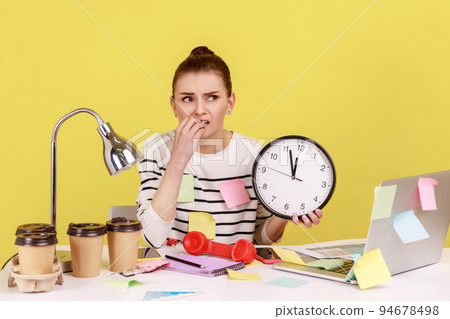 Nervous unhappy woman office worker biting nails holding big wall clock, sitting at workplace all covered with sticky notes, deadline. Indoor studio studio shot isolated on yellow background. Nervous unhappy woman office worker biting nails holding big wall clock, sitting at workplace all covered with sticky notes, deadline. Indoor studio studio shot isolated on yellow background. 94678498