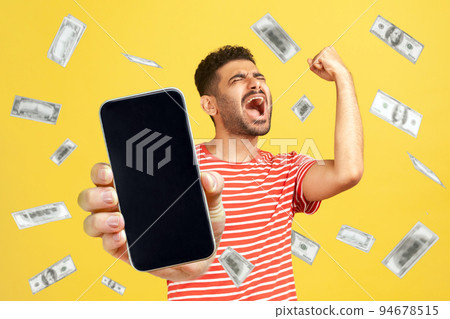 Money rain, winner and rich. Portrait of rejoicing happy bearded man wearing red T-shirt standing with smart phone in hands, showing blank screen. Indoor studio shot isolated on yellow background. 94678515
