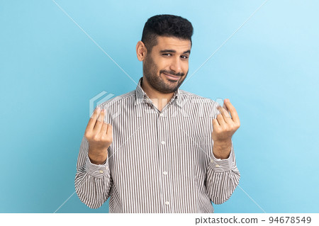 Cunning mercantile businessman with beard rubbing fingers showing money gesture, asking for salary, demanding bribe, wearing striped shirt. Indoor studio shot isolated on blue background. 94678549
