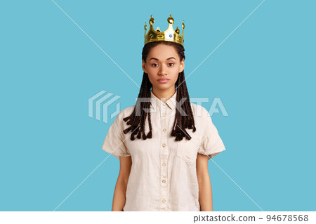Arrogant selfish woman with black dreadlocks egoistically looking at camera, posing with crown on head, pretending to be queen, wearing white shirt. Indoor studio shot isolated on blue background. 94678568
