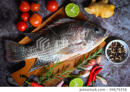 Tilapia with spice rosemary tomato lemon lime ginger garlic pepper chili on dark background, Fresh raw tilapia fish from the tilapia farm Tilapia with spice rosemary tomato lemon lime ginger garlic pepper chili on dark background, Fresh raw tilapia fish from the tilapia farm 94679356