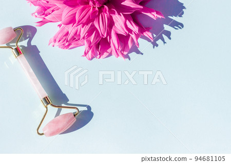 Beautiful pink flower and crystal rose quartz facial roller on blue background with copy space. Facial anti-age massage. Concept youth, beauty and self-care Beautiful pink flower and crystal rose quartz facial roller on blue background with copy space. Facial anti-age massage. Concept youth, beauty and self-care 94681105