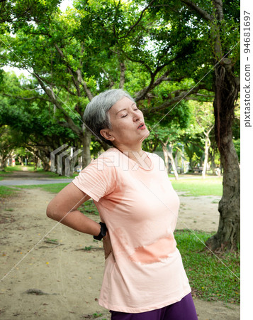 A 60-year-old Asian mature woman in an outdoor park caressing her lumbar area and living with back pain or back pain in her lower back. 94681697