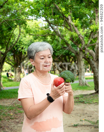 Portrait of happy senior adult elderly asia woman smiling standing and holding fresh vegetable and apple in the park. 94682309