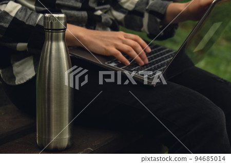 Water bottle. Reusable steel thermo water bottle on wooden bench. Sustainable lifestyle. Plastic free zero waste free living. Go green Woman student work study with laptop in park  94685014