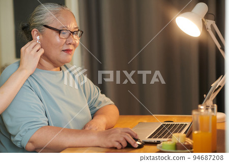 Grandson helping grandmother to put in earbuds when she is working on laptop Grandson helping grandmother to put in earbuds when she is working on laptop 94687208
