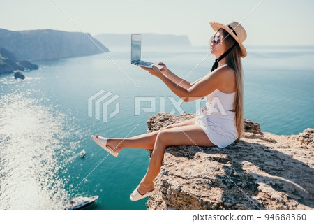Digital nomad, woman in the hat, a business woman with a laptop sits on the rocks by the sea during sunset, makes a business transaction online from a distance. Freelance, remote work on vacation. 94688360