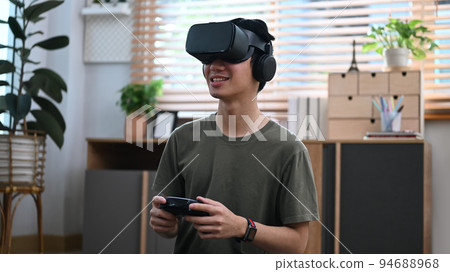 Smiling asian man holding joystick and playing video game with virtual reality glasses at home 94688968