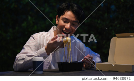 Happy young asian man eating take away noodles at a table at night market. Night life concept 94688971