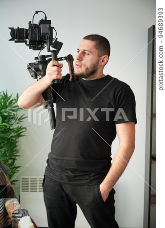 Portrait of videographer man posing indoors with camera mounted on gimbal stabilizer equipment. 94689393