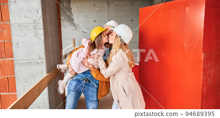 Woman kissing daughter while standing at construction site. Happy parents with child posing in apartment building under construction. Family life and new home purchase concept. Woman kissing daughter while standing at construction site. Happy parents with child posing in apartment building under construction. Family life and new home purchase concept. 94689395