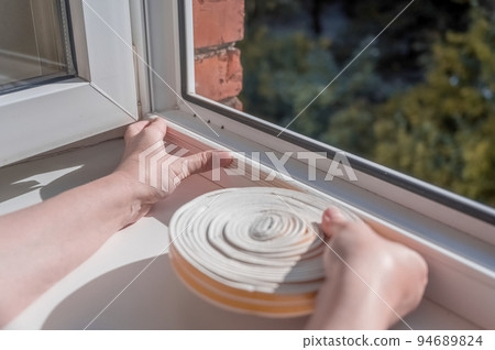 a woman glues a sealing rubber tape on a window in a living room. 94689824