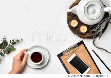 Young woman right hand writing on blank notebook on white table with tea cup, smartphone, and kettle beside in morning time Young woman right hand writing on blank notebook on white table with tea cup, smartphone, and kettle beside in morning time 94690084