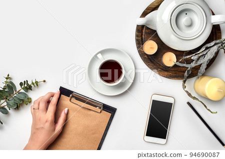 Young woman right hand writing on blank notebook on white table with tea cup, smartphone, and kettle beside in morning time Young woman right hand writing on blank notebook on white table with tea cup, smartphone, and kettle beside in morning time 94690087
