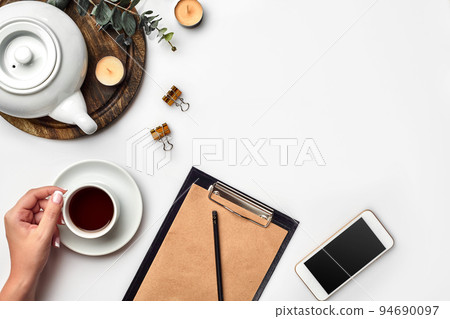 Young woman right hand writing on blank notebook on white table with tea cup, smartphone, and kettle beside in morning time 94690097