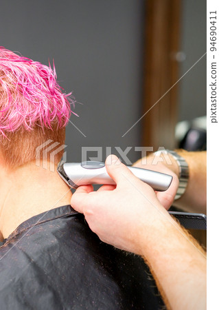 Male hairdresser shaves neck of a young caucasian woman with a short pink hairstyle by electric shaver in a hairdresser salon, close up 94690411