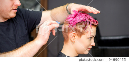 Haircut of dyed short pink wet hair of young caucasian woman by a male hairdresser in a barbershop Haircut of dyed short pink wet hair of young caucasian woman by a male hairdresser in a barbershop 94690455