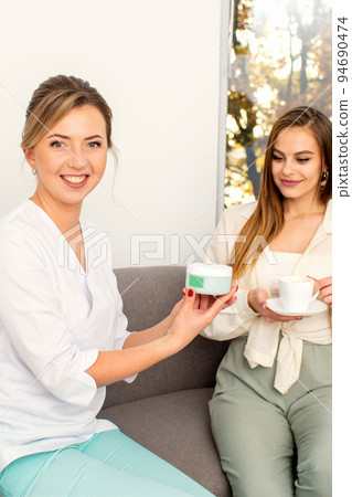 Beautician offering product for the young woman holding a white plastic jar with a cream sitting on the sofa 94690474
