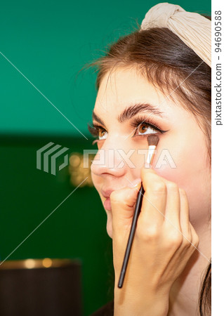 Make-up in the process. The hand of the makeup artist applies eye shadow under the eyes of a young beautiful caucasian model woman 94690588