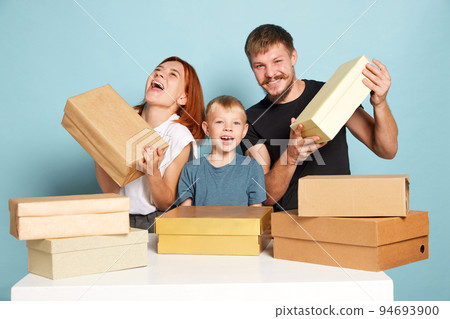Unpacking. Friendly family, parents and child open cardboard boxes, receive parcels unpack after online shopping over blue background. Black Friday concept Unpacking. Friendly family, parents and child open cardboard boxes, receive parcels unpack after online shopping over blue background. Black Friday concept 94693900