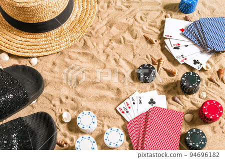 Beachpoker. Chips and cards on the sand. Around the seashells, hat and flip flops. Top view Beachpoker. Chips and cards on the sand. Around the seashells, hat and flip flops. Top view 94696182