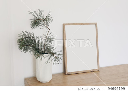 Christmas Scandinavian interior. Minimal winter artistic composition. Blank vertical wooden picture frame mockup. Pine tree branches in vase on table, desk. Beige wall background. Empty copy space. 94696199