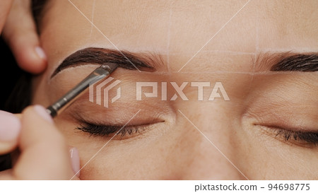 Woman is  on the Eyebrow coloring procedure. Professional lamination procedures of female eyebrows in beauty salon. Closeup view of client face and stylist's hands working.  Beauty care concept. 94698775