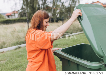 Young happy woman putting garbage in green trash bin on back yard background. Environmental conservation saving planet from contamination. Plastic pollution problem Young happy woman putting garbage in green trash bin on back yard background. Environmental conservation saving planet from contamination. Plastic pollution problem 94699721
